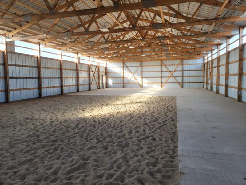 Large indoor arena with sand and concrete floors, wooden beams, and sunlight through windows.