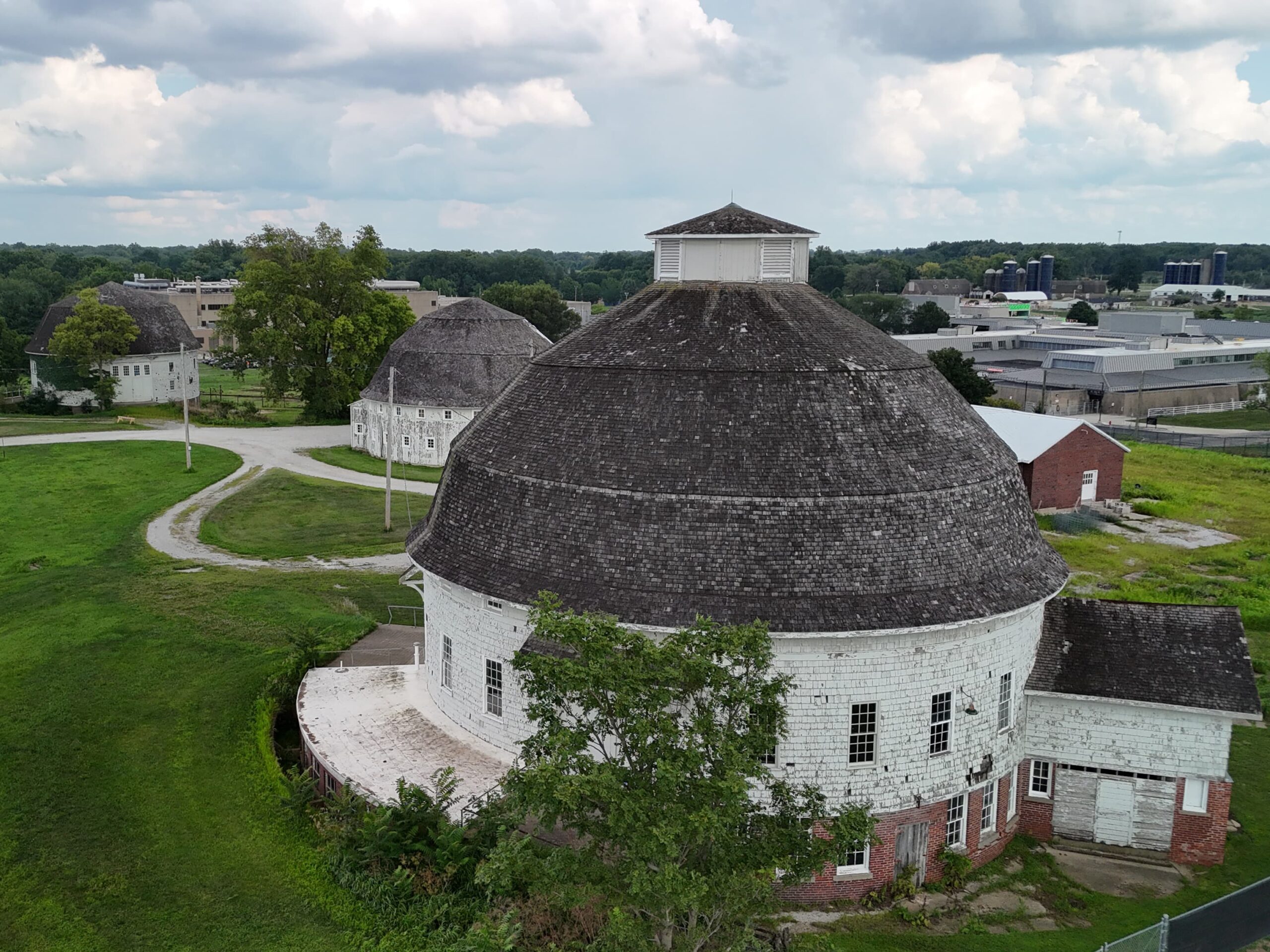 Aerial view of round, weathered farm buildings with shingled roofs, situated on lush green grass, under a partly cloudy sky. Peaceful rural scene.