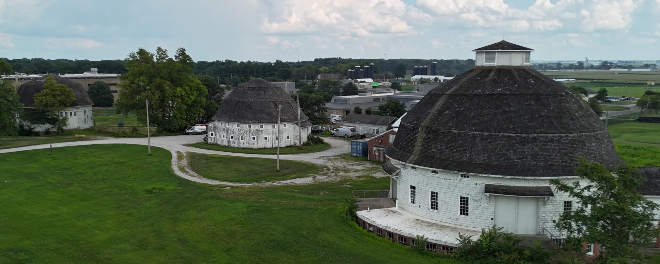 Aerial view of two white round barns with dark roofs on lush green grounds, surrounded by trees and distant buildings under a cloudy sky. Tranquil scene.