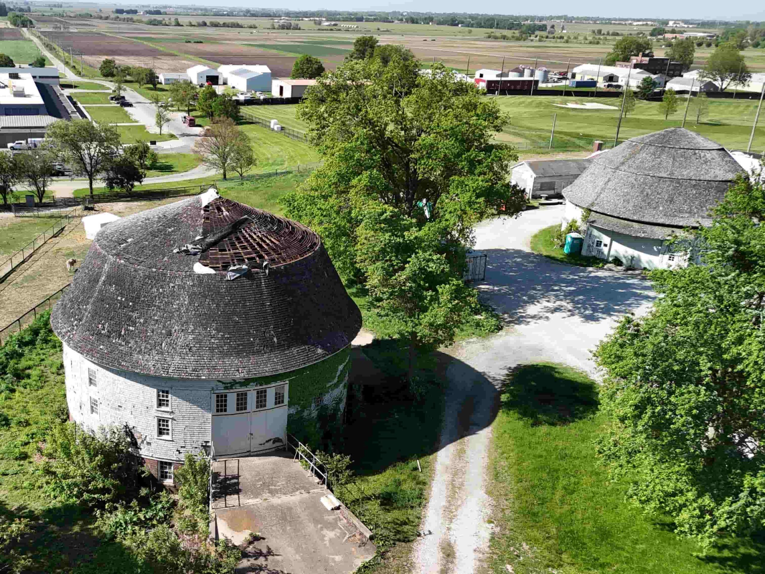 Aerial view of two round barns, including one with a partially damaged roof, surrounded by trees and farmland.