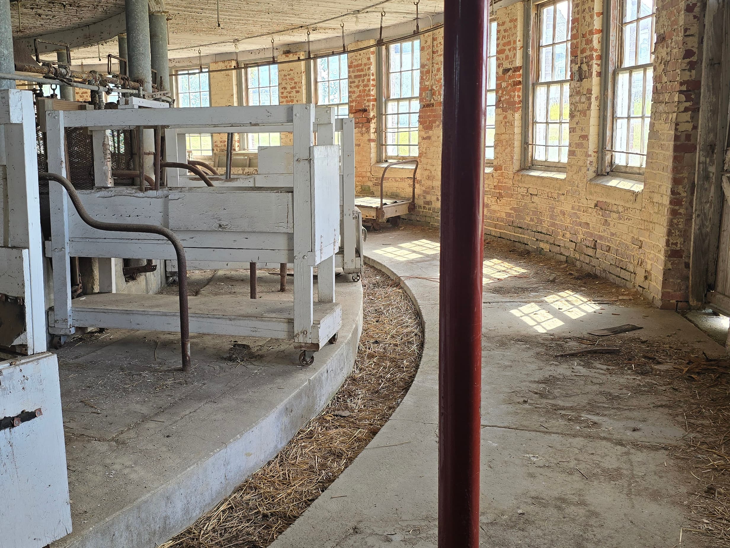 Interior of an old circular barn with brick walls, windows, a white wooden structure, and a concrete floor.
