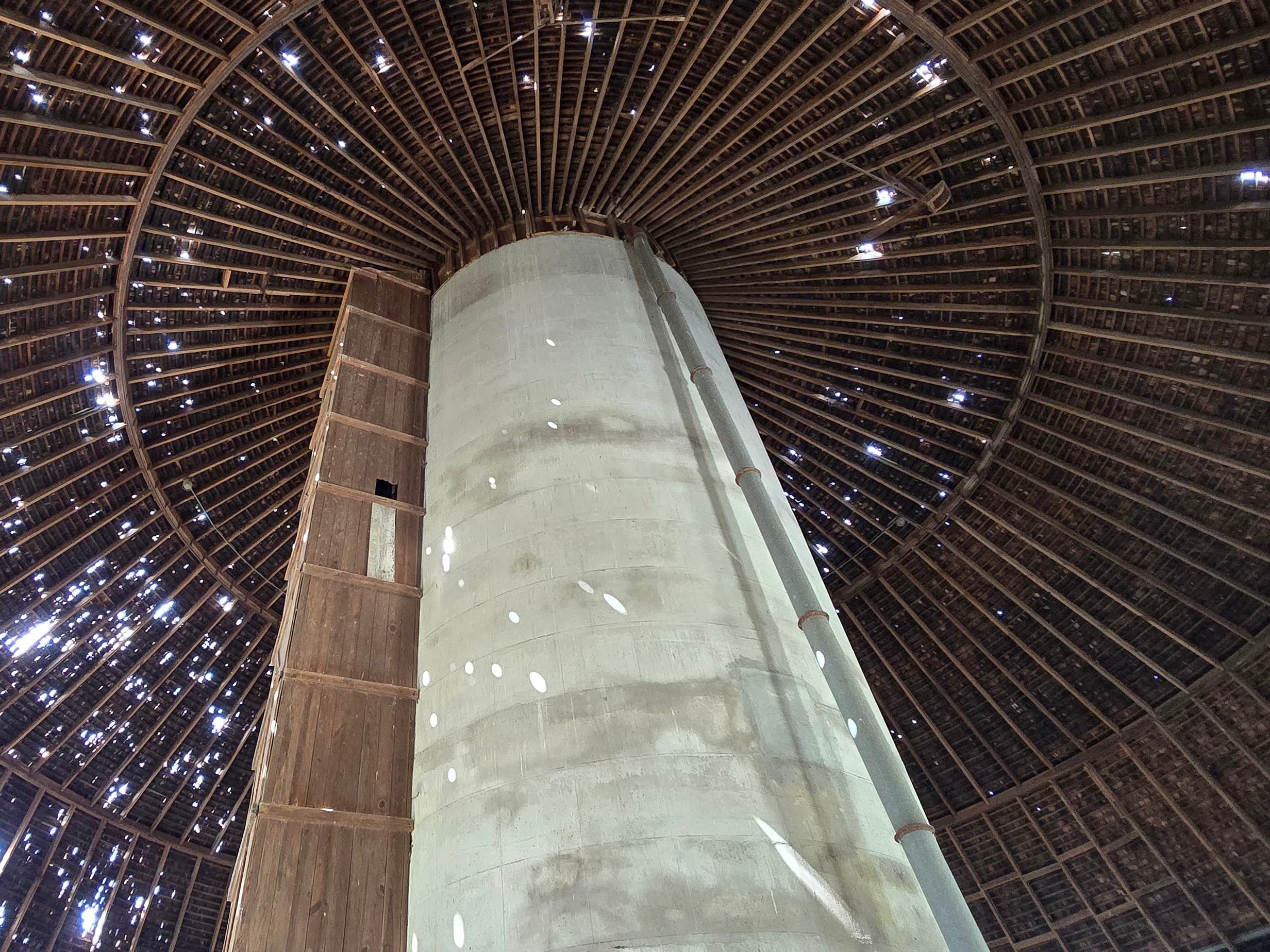 Interior view of a domed wooden roof with a tall concrete cylinder in the center, light filtering through gaps in the beams.