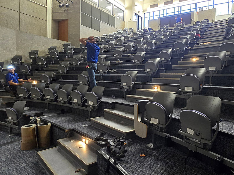 Workers install new tiered seating in LAC 100 lecture hall, with tools and materials visible during renovation.