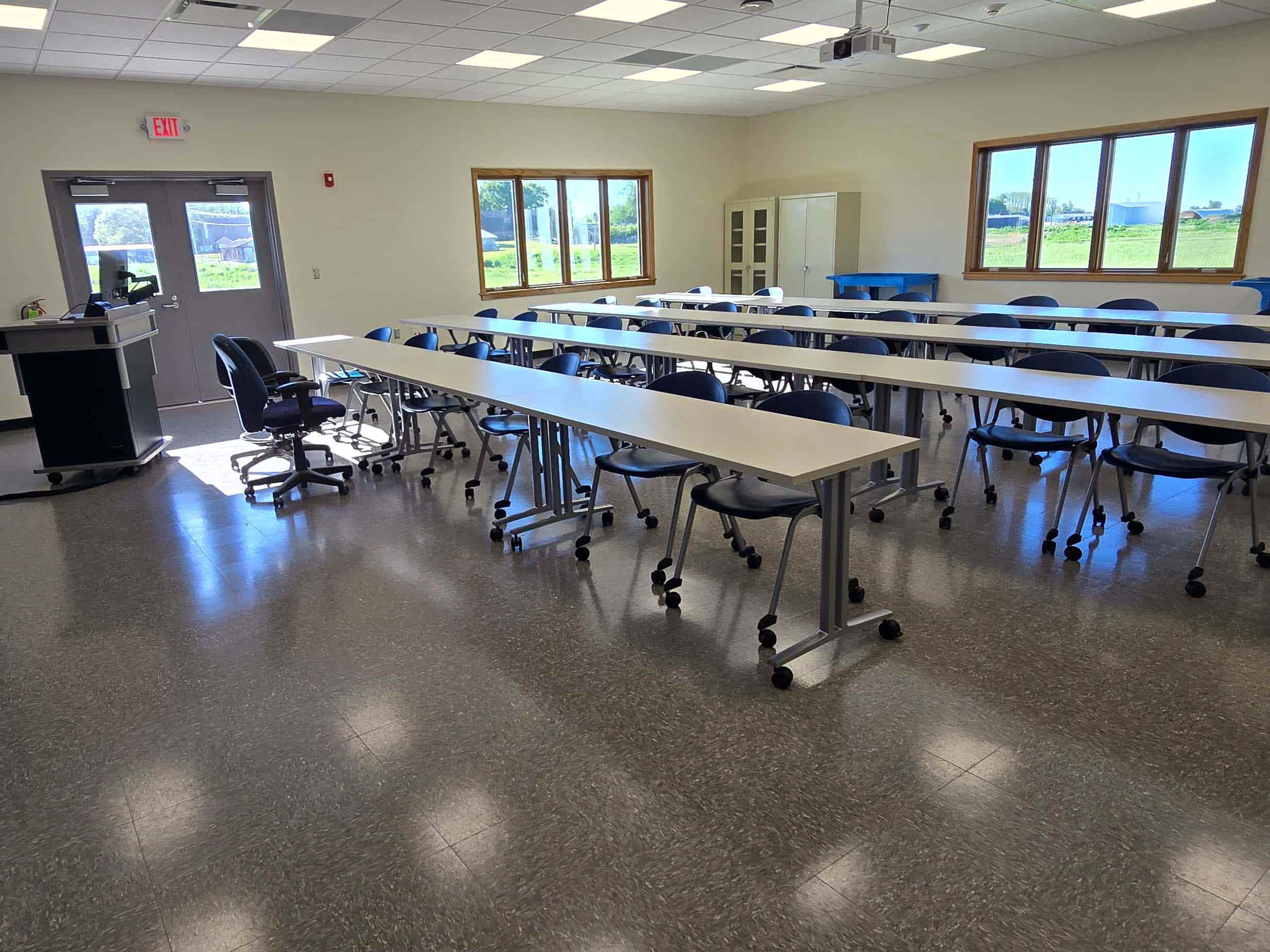 Classroom with rows of tables, rolling chairs, and a lectern near large windows.
