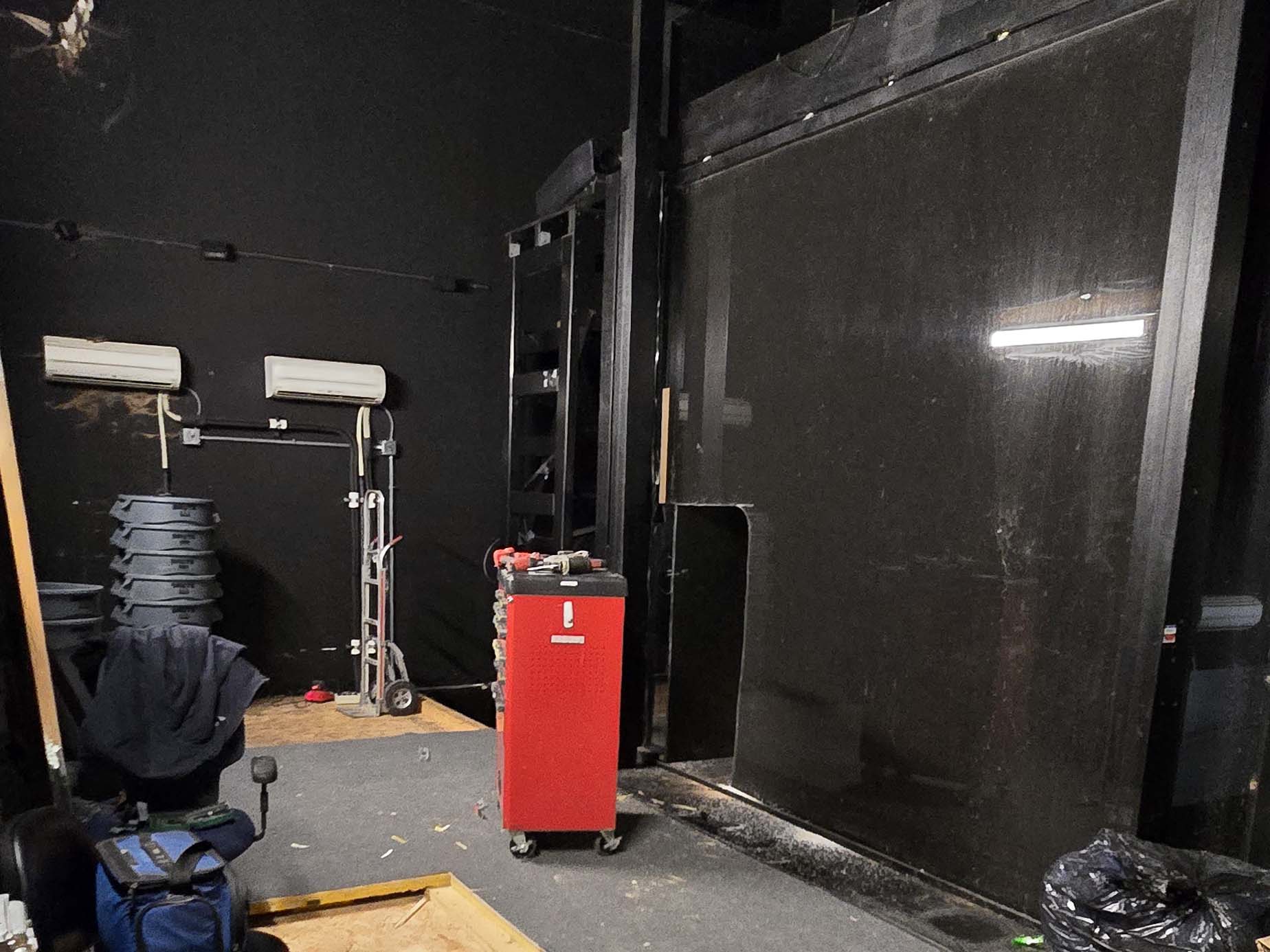 Interior view of an unfinished room with black walls, a large black panel, stacked chairs, a red tool chest on wheels, and miscellaneous equipment, indicating a space under renovation.
