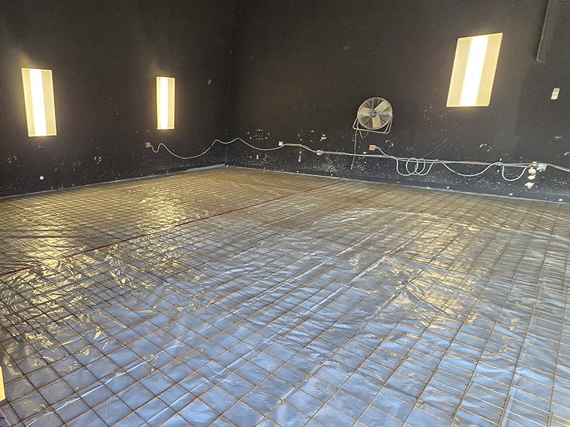 Interior view of a room under construction with black walls and three rectangular light panels. The floor is covered with plastic sheeting and a grid of rebar, indicating preparation for concrete pouring. Electrical wiring and a wall-mounted fan are visible along the back wall.