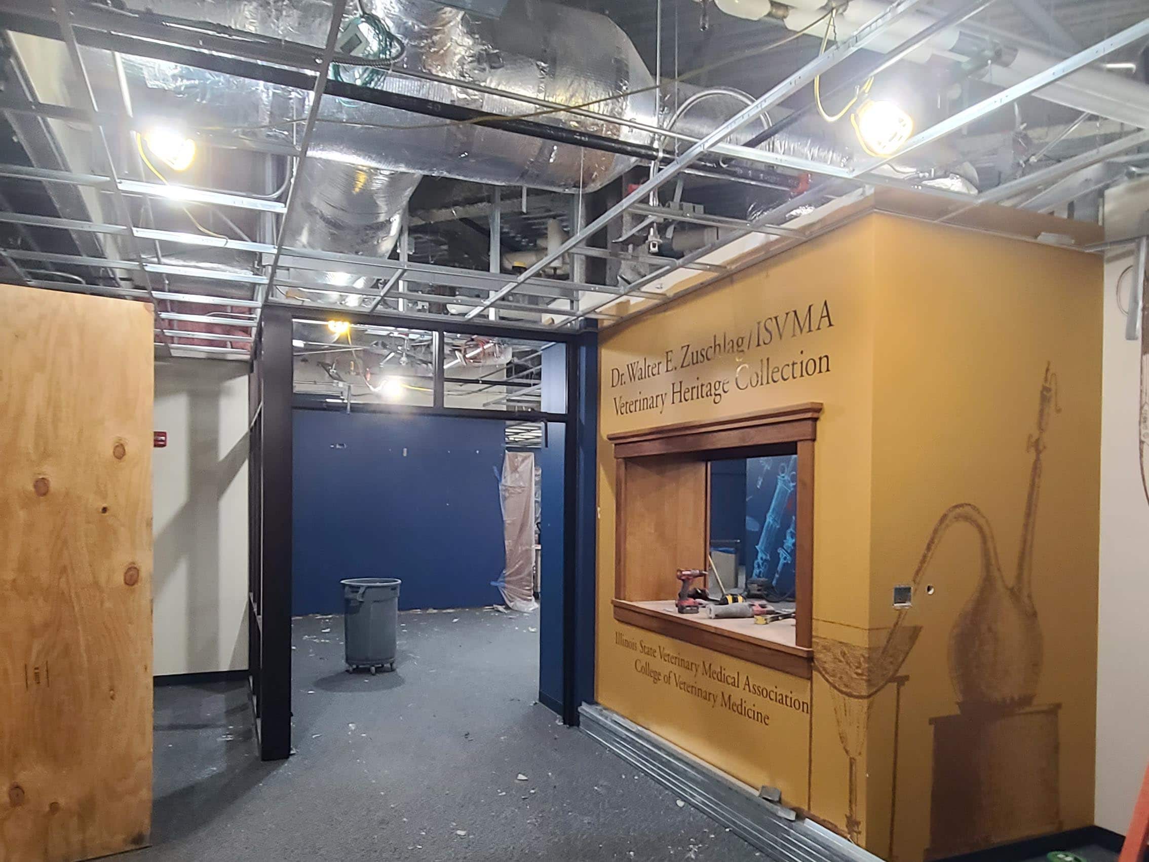 Interior hallway under renovation with exposed ceiling ducts, construction materials, and a veterinary heritage display wall.