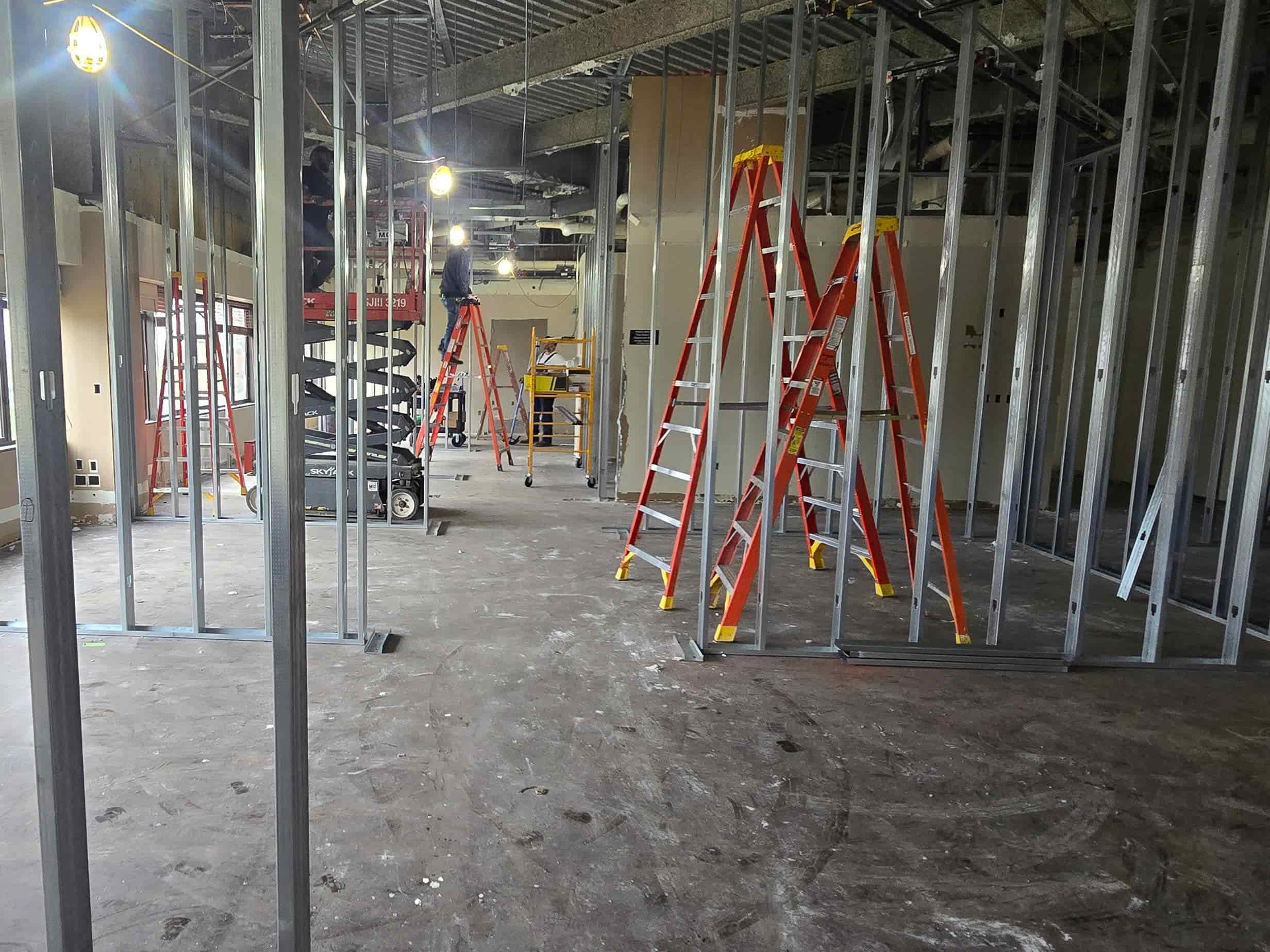 Interior renovation area with new metal wall framing, ladders, work lights, and construction equipment.