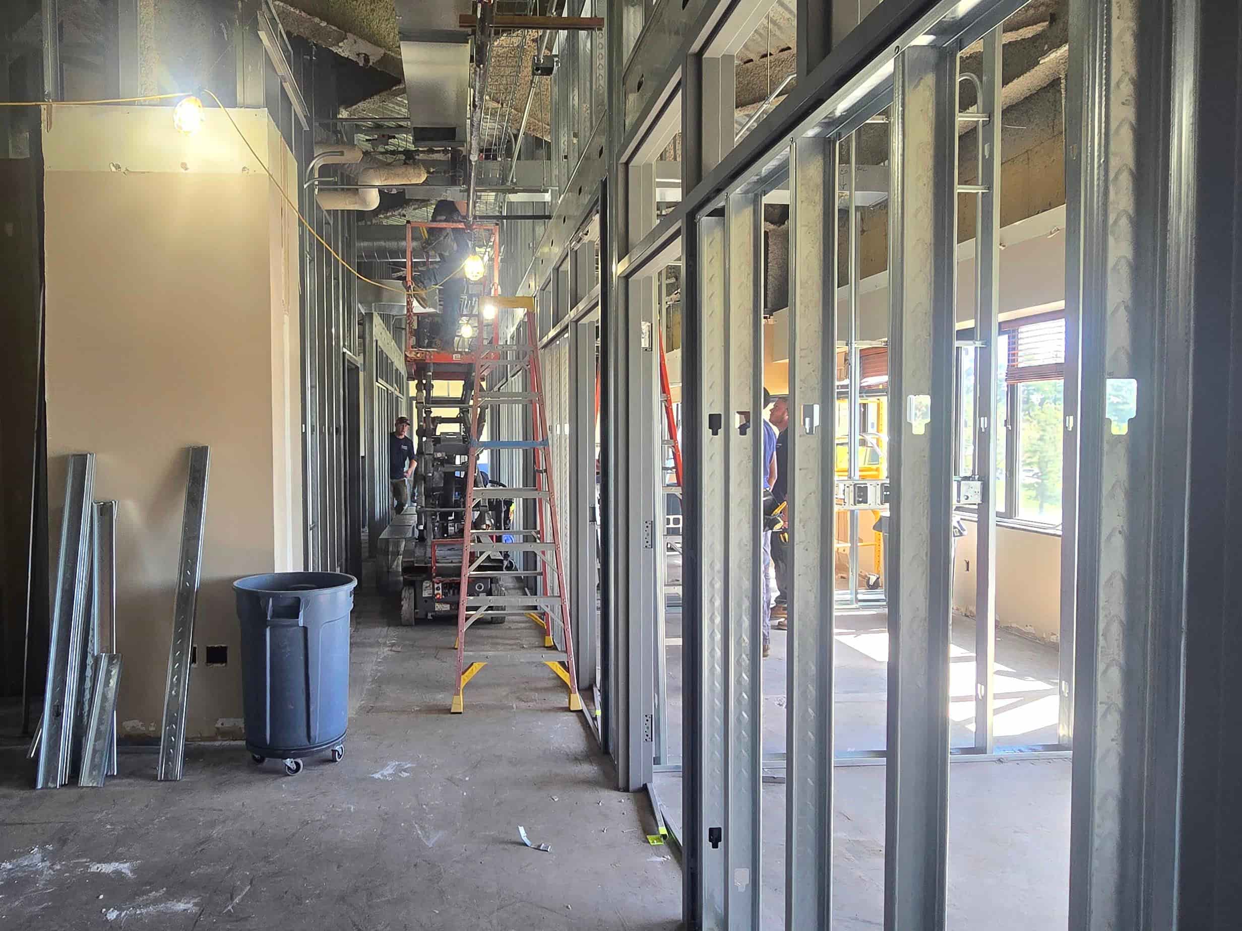 Interior renovation corridor with new metal wall framing, ladders, a scissor lift, and construction activity.