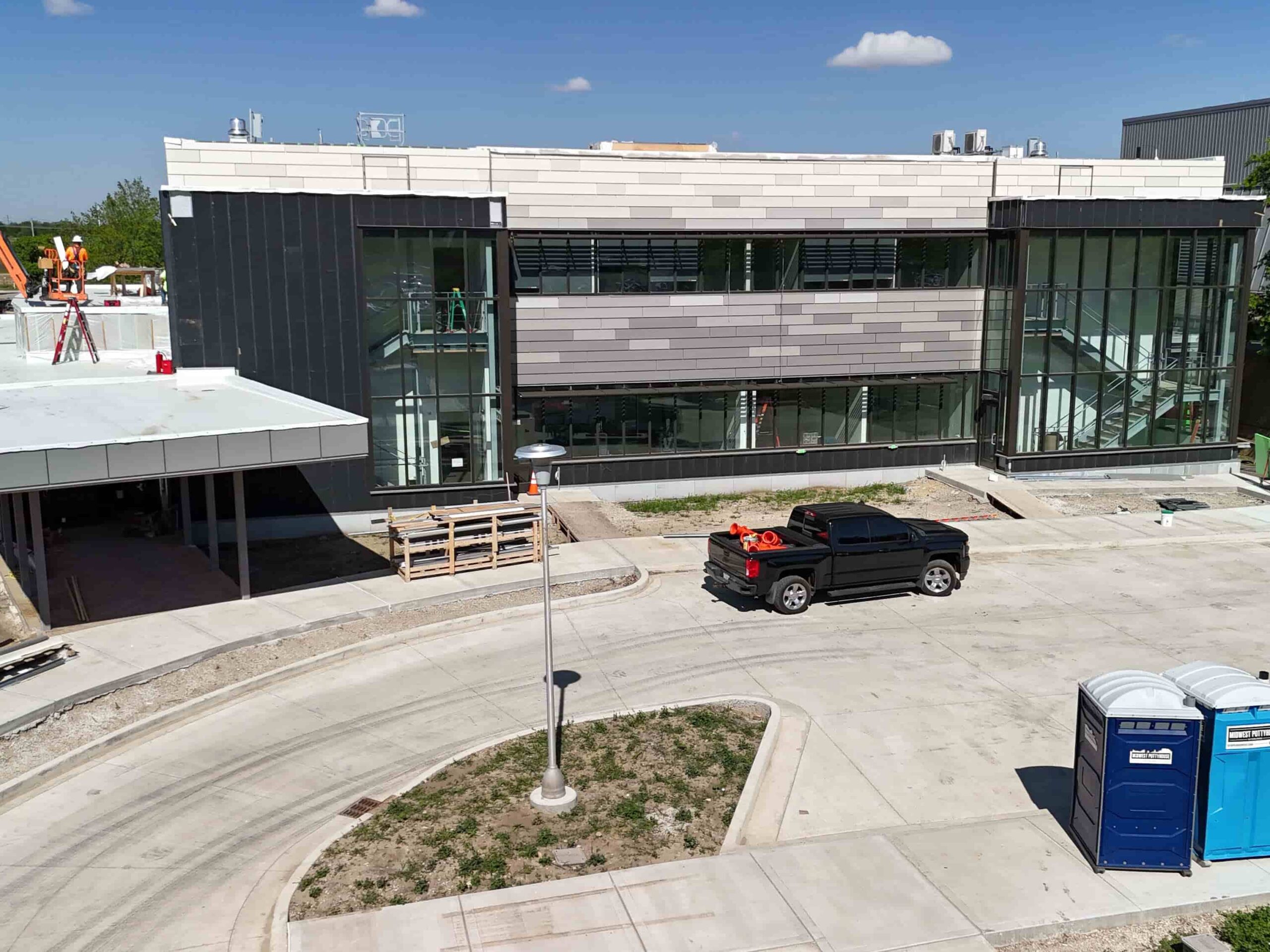 Two-story building under construction with glass windows, workers on the roof, a pickup truck, and portable toilets.