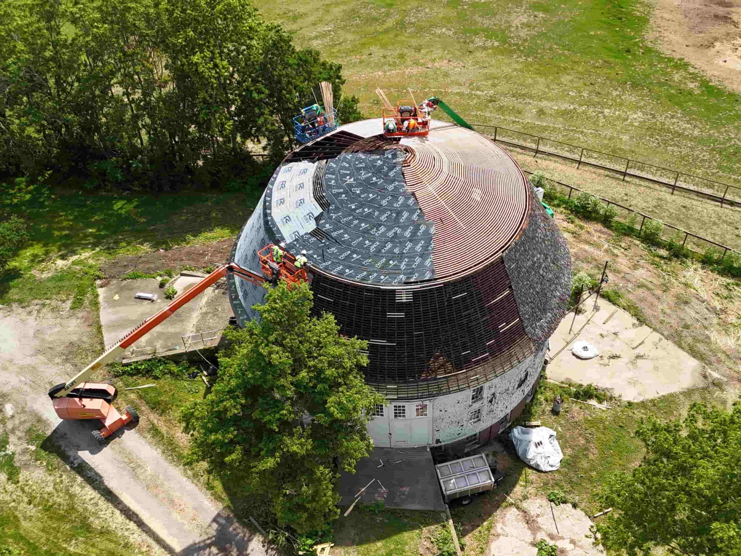Aerial view of the University of Illinois Round Barn with workers repairing the circular roof using lifts and safety equipment.