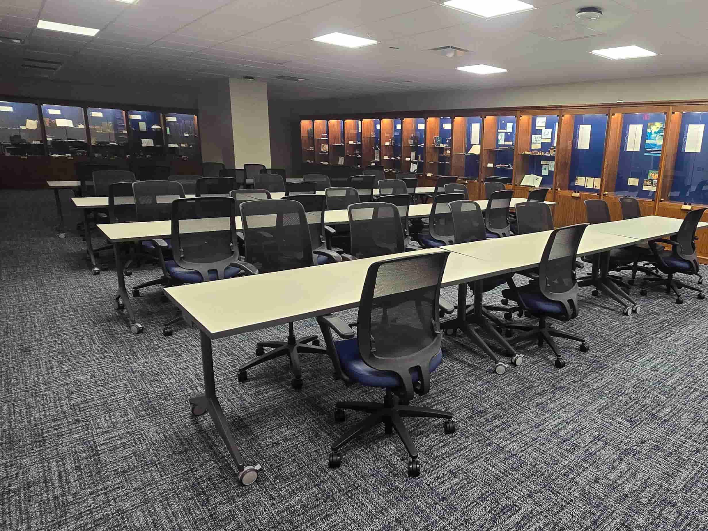 Modern classroom or training room with rows of tables and rolling chairs, surrounded by illuminated display cabinets.