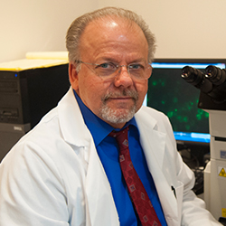 Raymond Rowland in a lab coat, sitting next to a microscope, looking at the camera