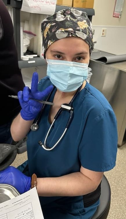 Our clinic intern, Sofia, is sitting on a stool with a pen in her hand and stethoscope around her neck, dressed in scrubs with a surgical cap and facial mask on. She is holding her right hand up and showing a peace sign or the pointer and middle finger are making a "V" shape.
