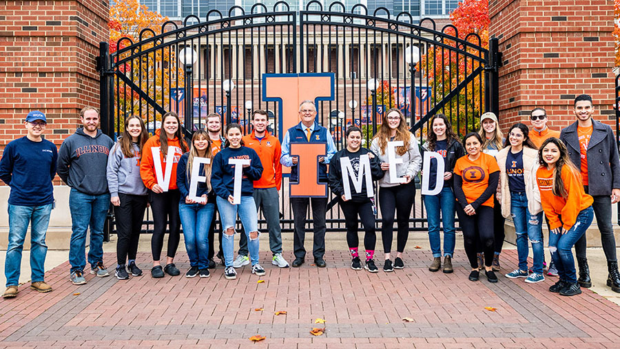 Dean Constable poses with a dozen or so vet students in front of gate with large Block I logo