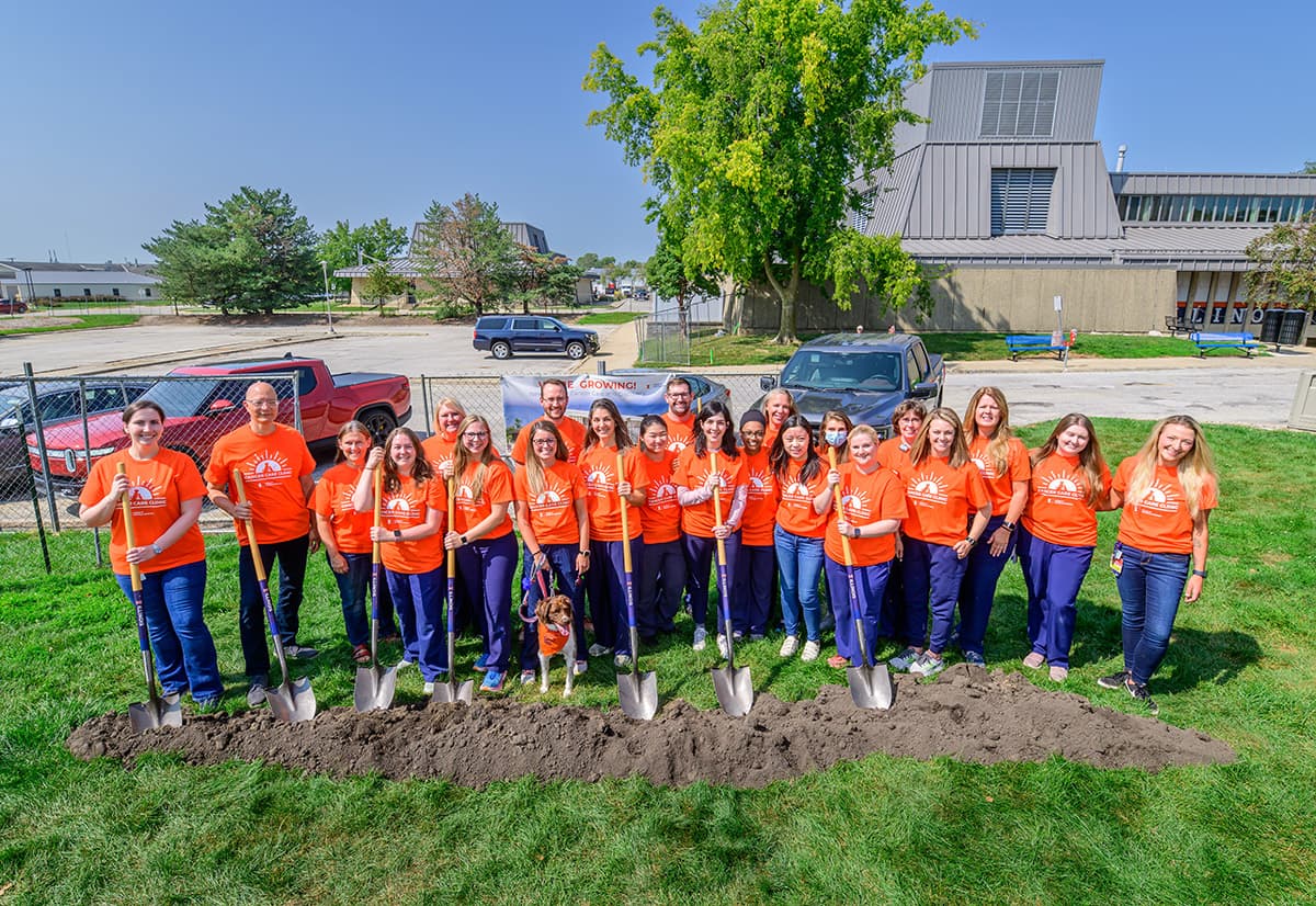 The oncology team at Vet Med lined up in orange t-shirts with seven clinicians holding Illinois-branded shovels behind a small pile of dirt for a groundbreaking photoshoot in celebration of the new oncology wing