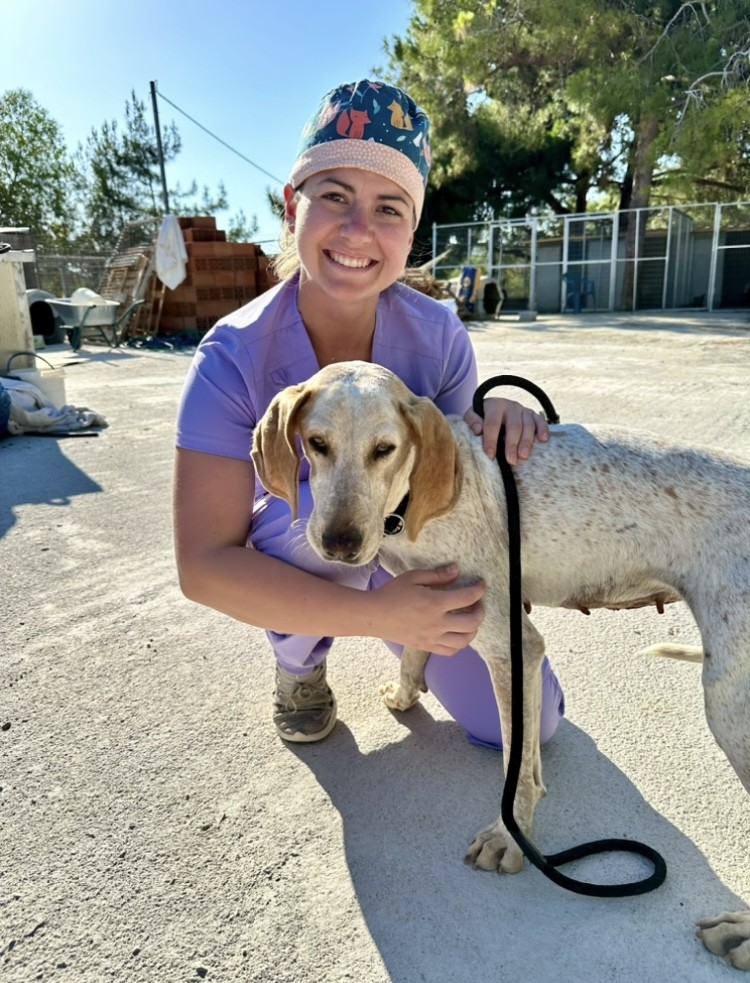 A veterinary student in purple scrubs smiling with a beige-colored dog.