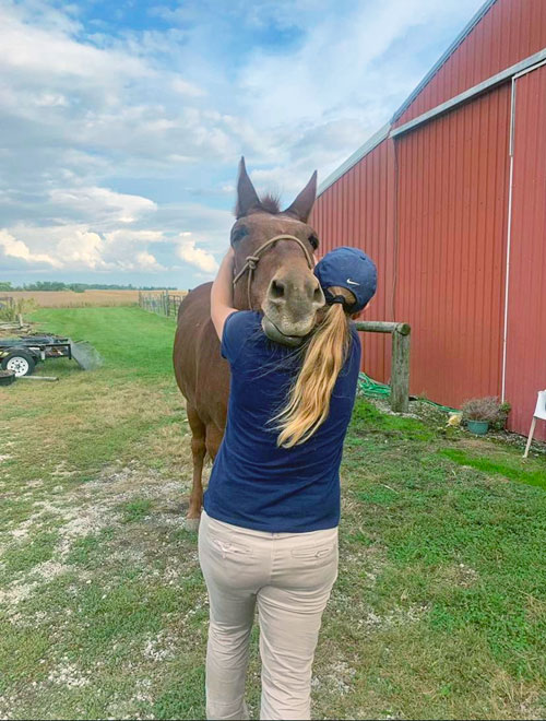 A woman with a long ponytail in a ball cap appears to be hugging a mule. This is Dr. Foreman-Hesterberg performing a chiropractic adjustment on Art the mule.