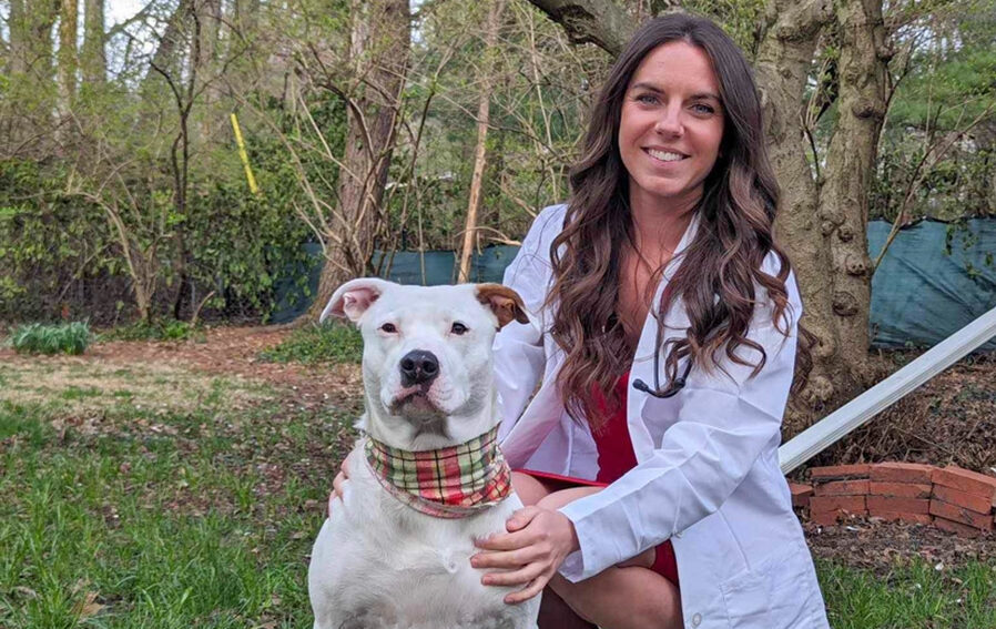 Gina Kowalewicz wearing a white veterinary coat and posing with her dog outdoors.
