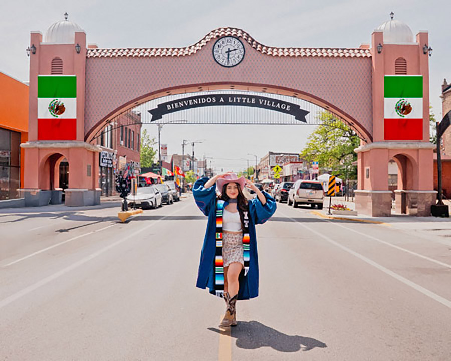 Naomi Esquivel posing in her graduation gown with a cowboy hat in front of an archway that reads "Bienvenidos a Little Village."