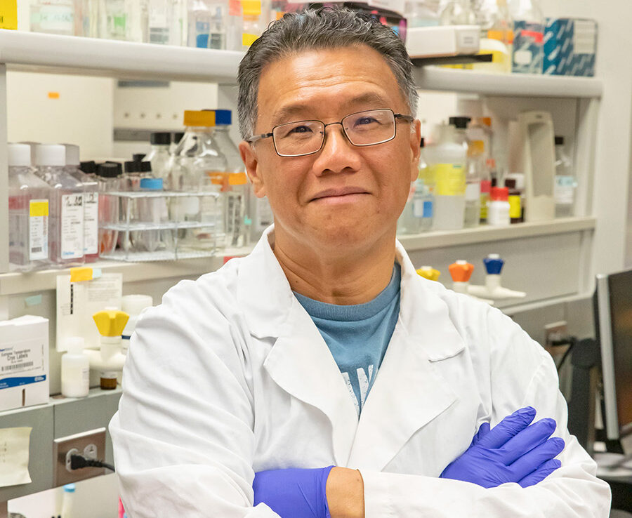 Gee Lau posing with arms crossed in a white lab coat and purple gloves in a clinical setting.