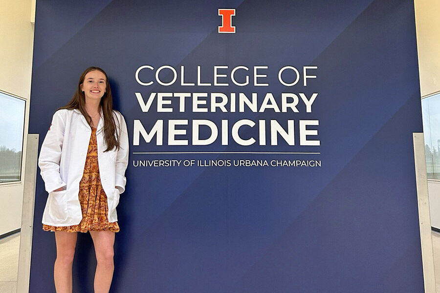Annie Marlowe in her white coat in front of a blue wall inside the University of Illinois College of Veterinary Medicine.