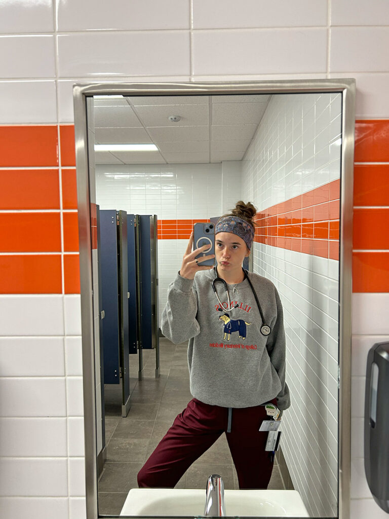 Annie Marlowe taking a mirror selfie in scrubs in a white and orange tiled bathroom.