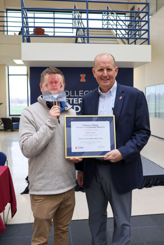 Jake Bough holding a cardboard cut out of Randy Dunker's face to accept his college excellence award on his behalf