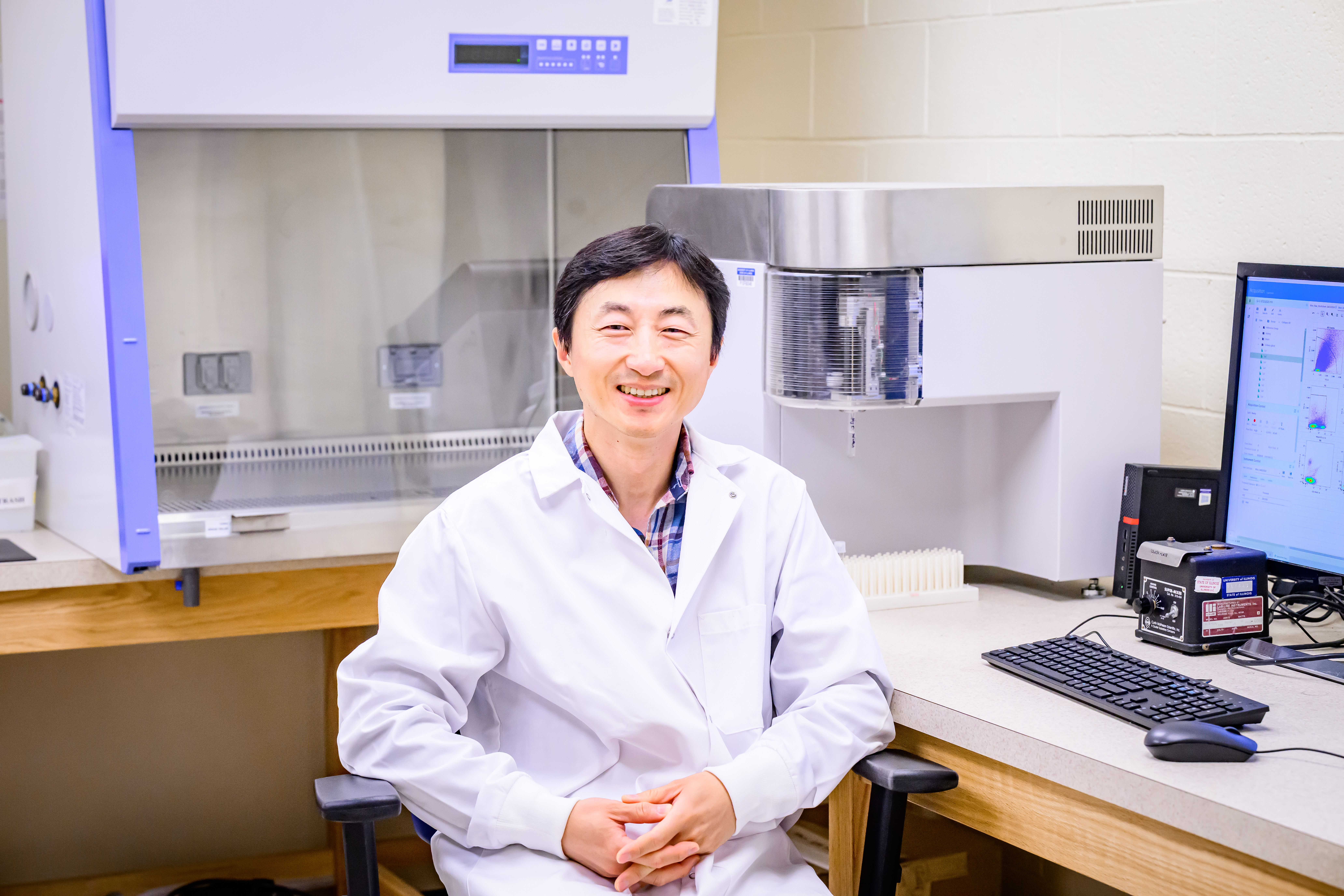 Dr. Taejoon Won sits at a desk in a research lab smiling at the camera white wearing a lab coat.